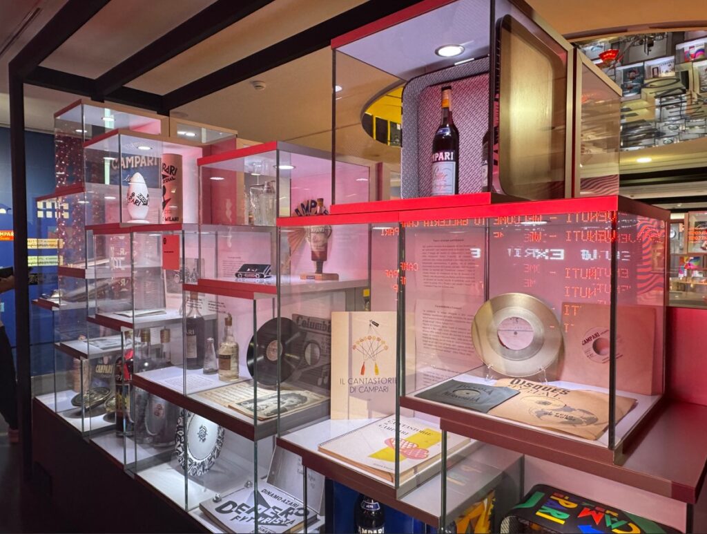 The display case showcases an array of Campari memorabilia, including bottles, records, and vintage advertisements, accented by a warm, pinkish light.