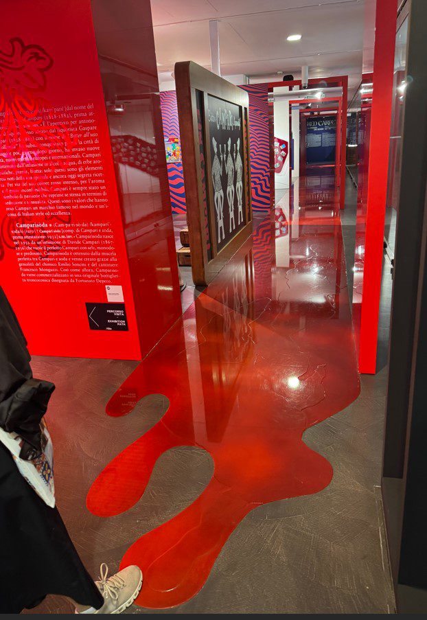 A vibrant museum hallway features glossy red liquid-like flooring, bold red walls with text, and a central art display of white figures.