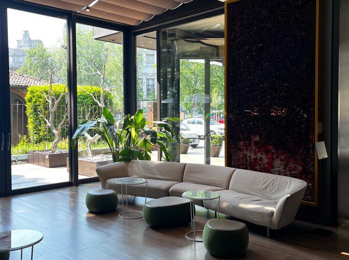 Modern lounge with beige curved sofa, green poufs, round tables, and large windows. Sunlight illuminates indoor plants and outdoor greenery.