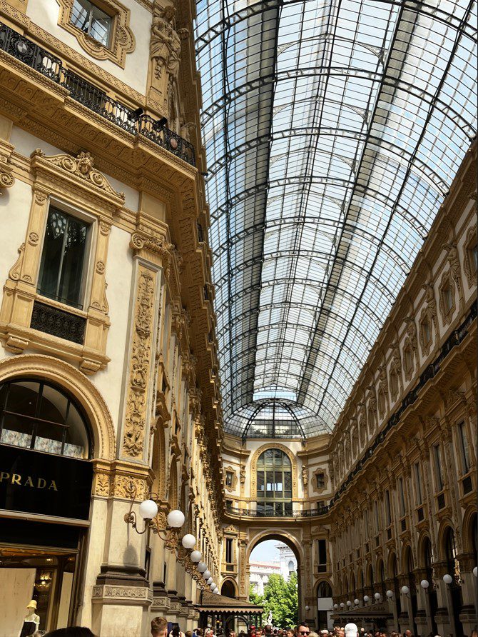 Ornate mall interior with arched glass ceiling, decorative columns, detailed moldings, and spherical lamps.